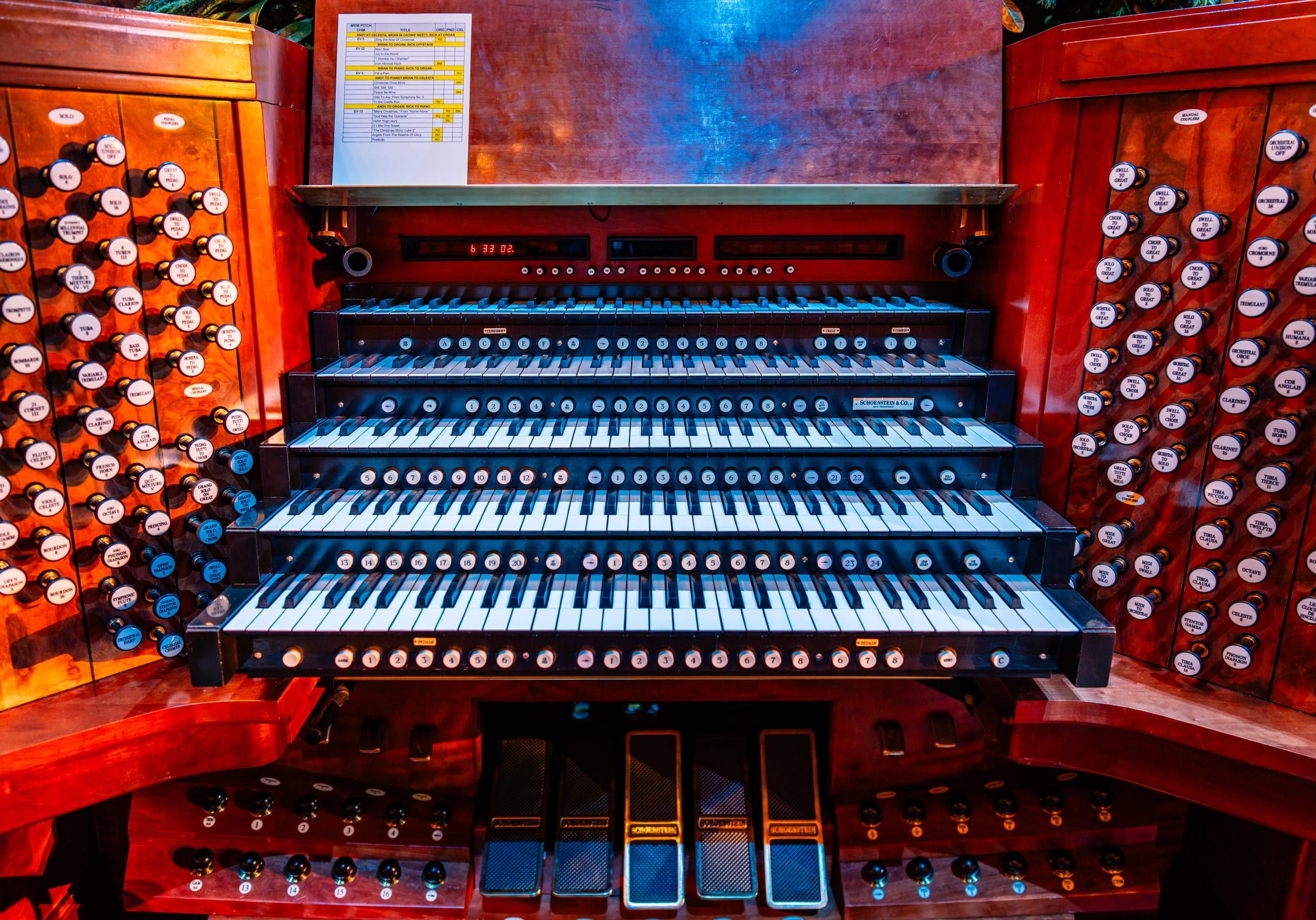 The five-manual console of a Schoenstein pipe organ, its banks of stop knobs lit in red and blue, with a performance set list taped above the music desk.