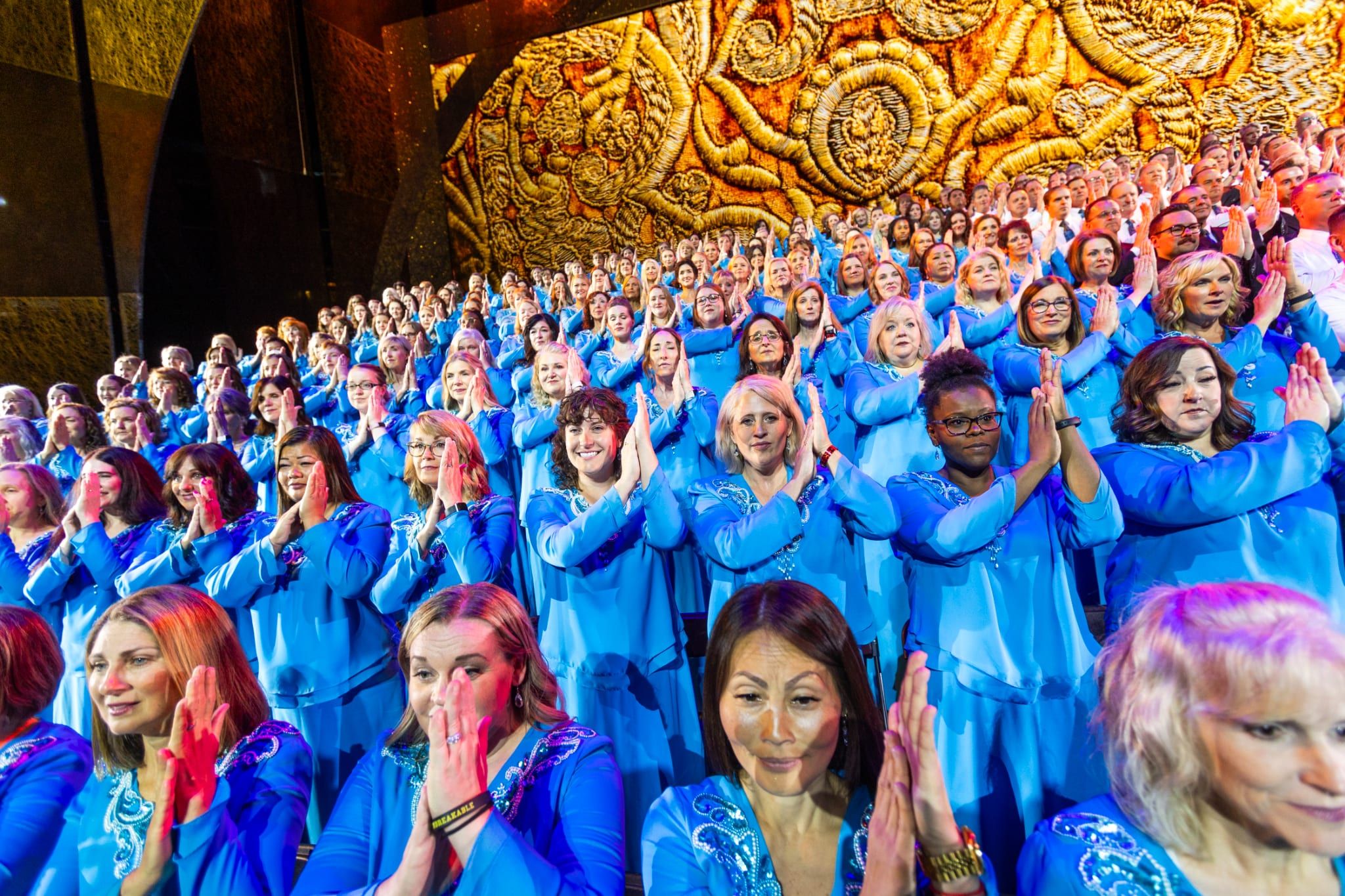 Choir members in bright blue robes applaud from the risers, a vast golden textured backdrop rising behind them.