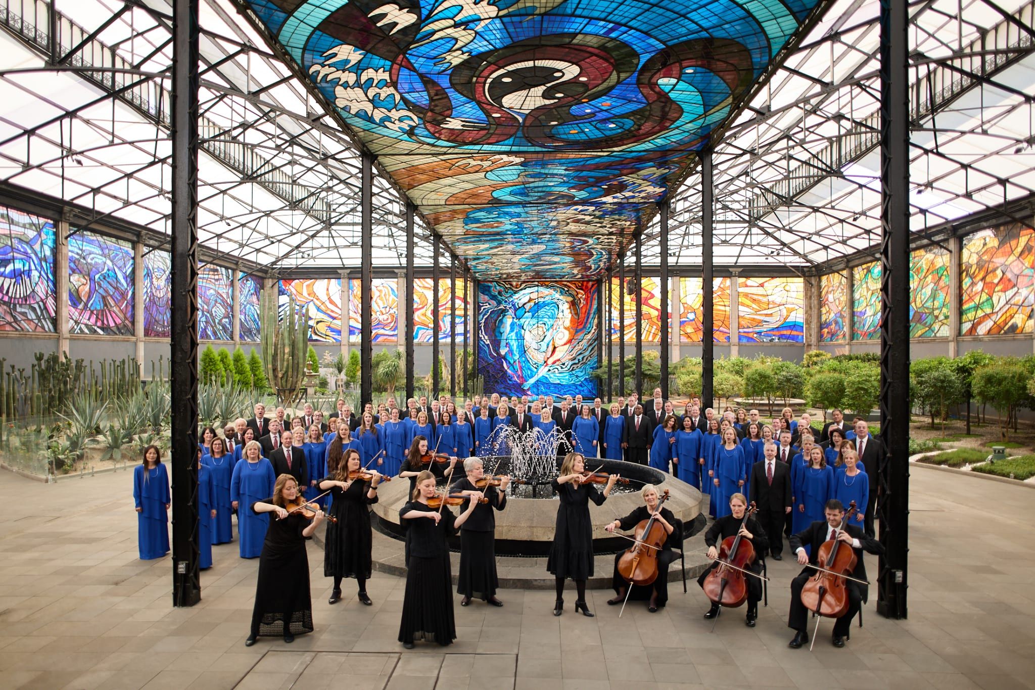 The Choir in blue robes and the Orchestra performing beneath a vast stained-glass ceiling, ringed by cacti and gardens.